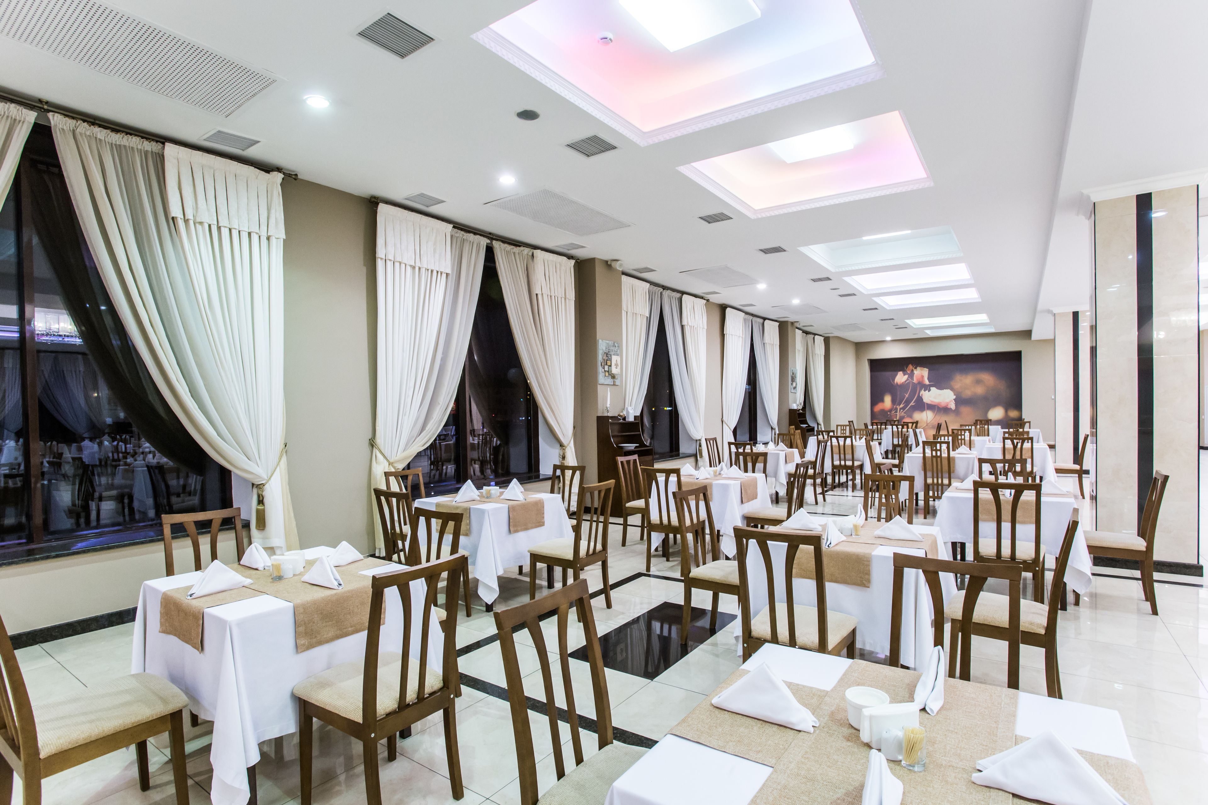 A spacious dining area with multiple tables and chairs, large windows with curtains, and a ceiling with skylights.