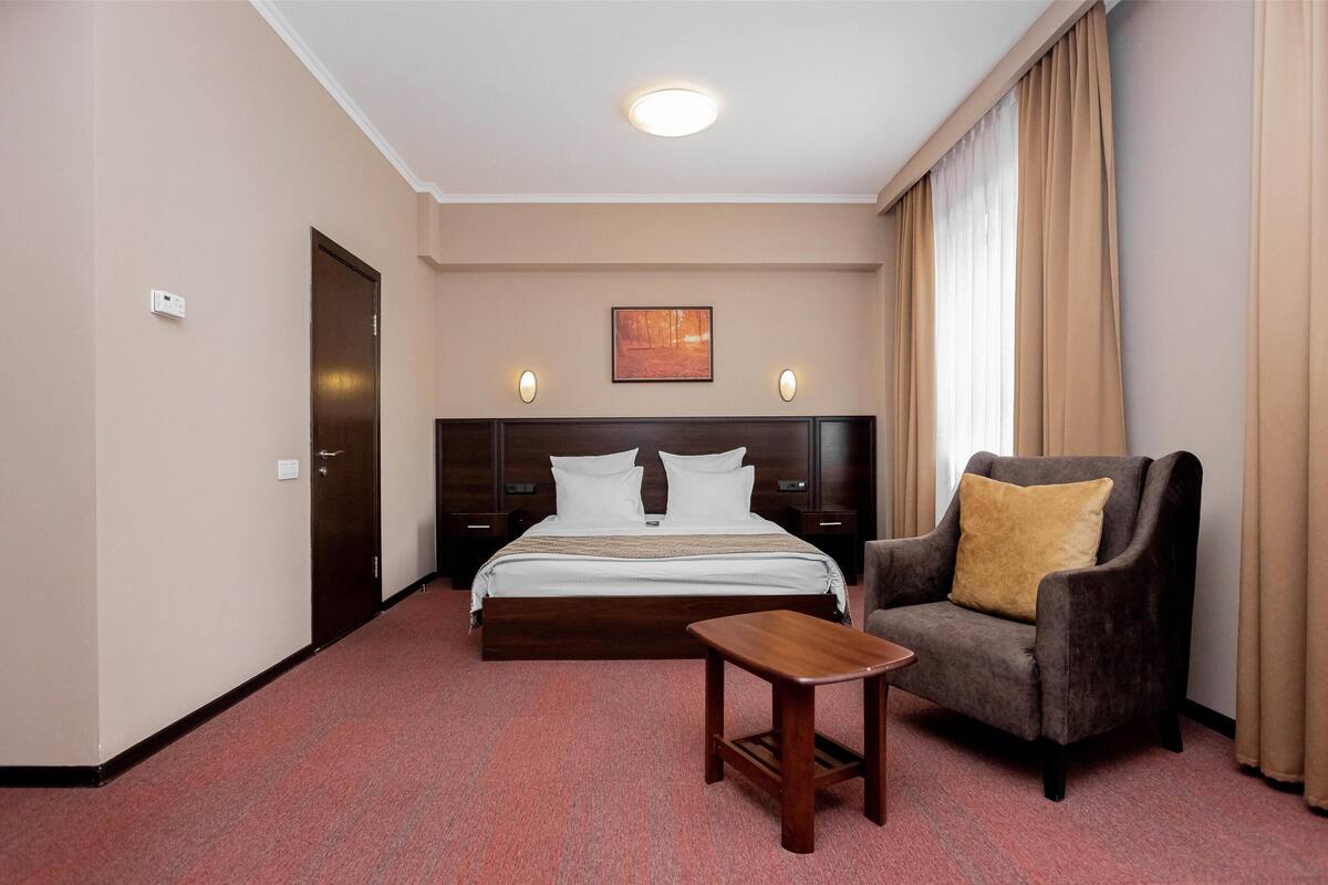 A hotel room with a large bed, a chair, a small table, and a painting on the wall.