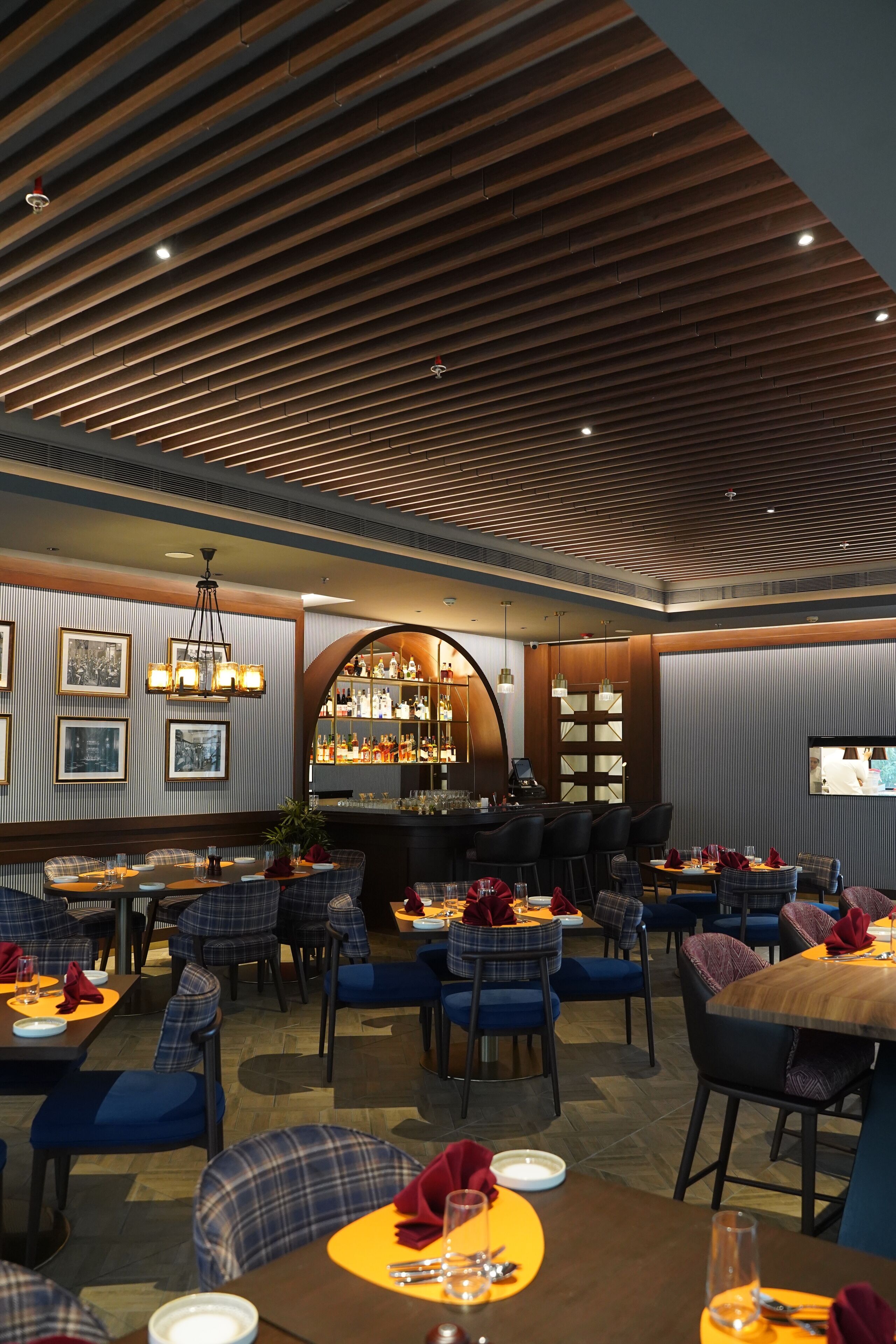 Un restaurant au décor moderne, avec des panneaux de bois au plafond, des chaises rembourrées en tissu bleu et un bar agrémenté d’un grand miroir et d’étagères remplies de bouteilles.