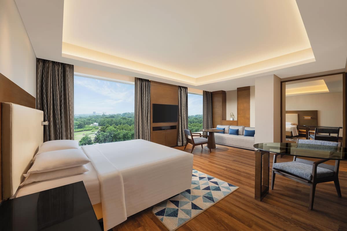 A modern hotel room with a bed, a desk, a sofa, and a large window offering a view of the outdoors.