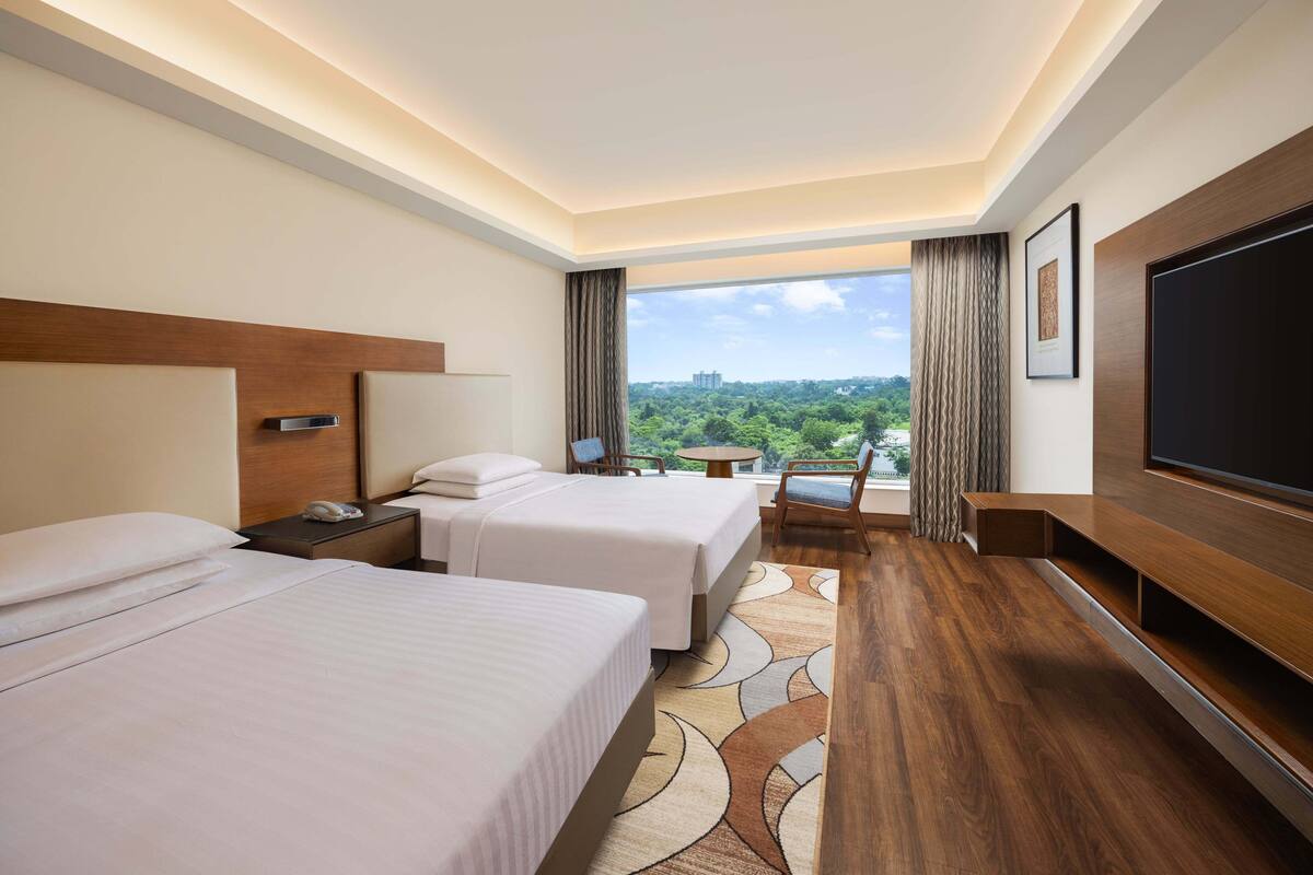 Habitación de hotel con dos camas, televisión, escritorio y vista a la ciudad.