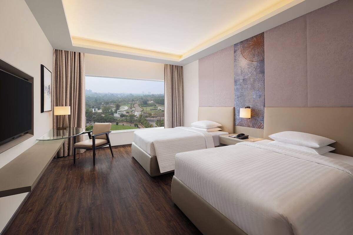Habitación de hotel con dos camas, un escritorio, una silla, televisión y vista a un paisaje urbano.