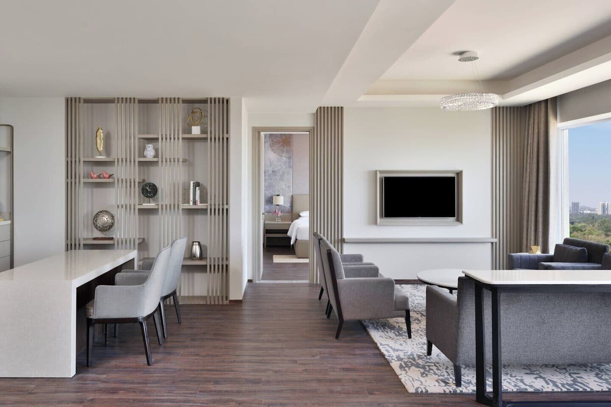A modern hotel room with a dining area, a TV mounted on the wall, and a view of the cityscape.