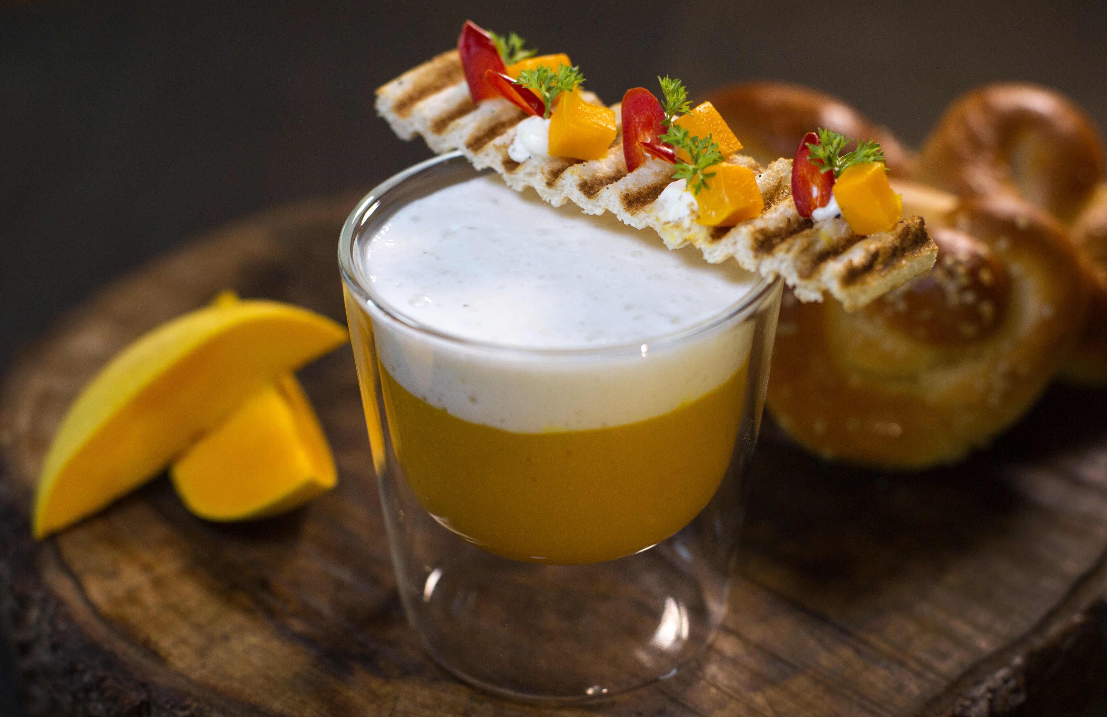 A glass of a layered beverage with a decorative topping, accompanied by a pretzel and mango slices on a wooden surface.