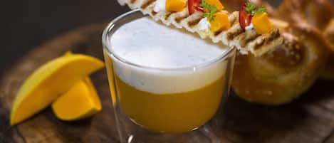 A glass of a layered beverage with a decorative topping, accompanied by a pretzel and mango slices on a wooden surface.