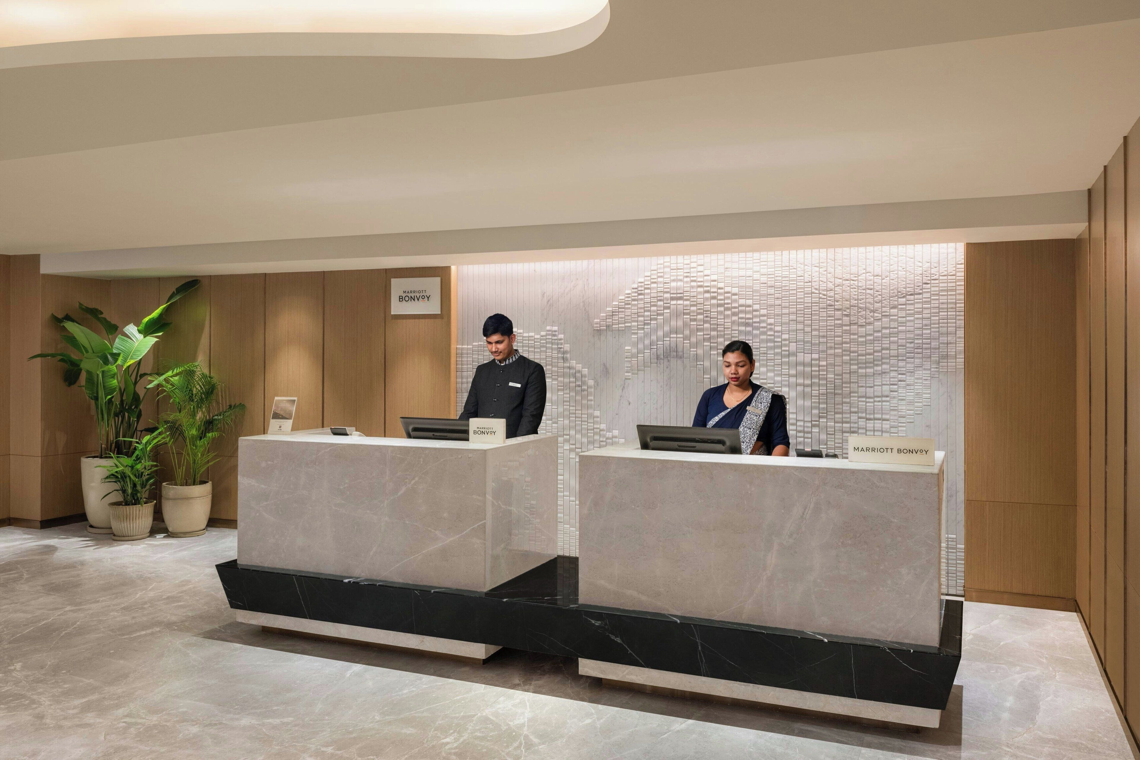 Área de recepção de hotel com dois recepcionistas em balcões de mármore, uma grande planta em vaso e um design moderno na parede.