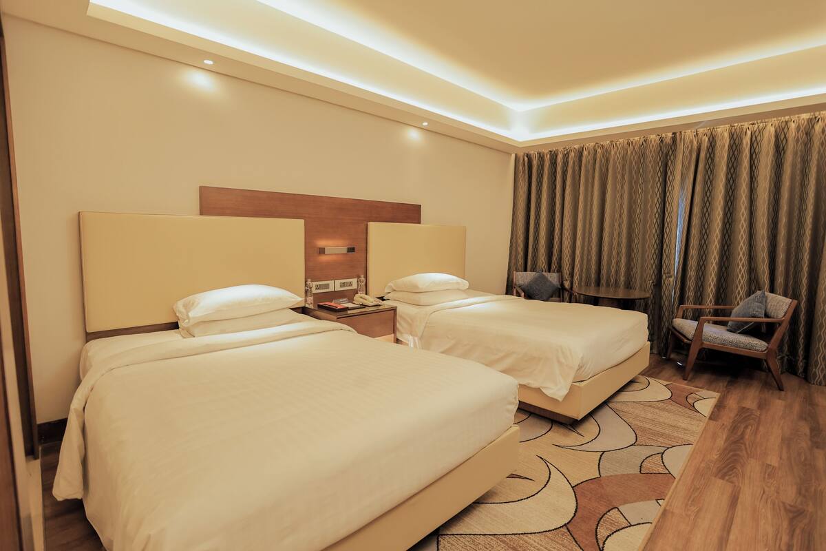 Habitación de hotel con dos camas, una mesita de noche, una silla y un ventanal con cortinas.