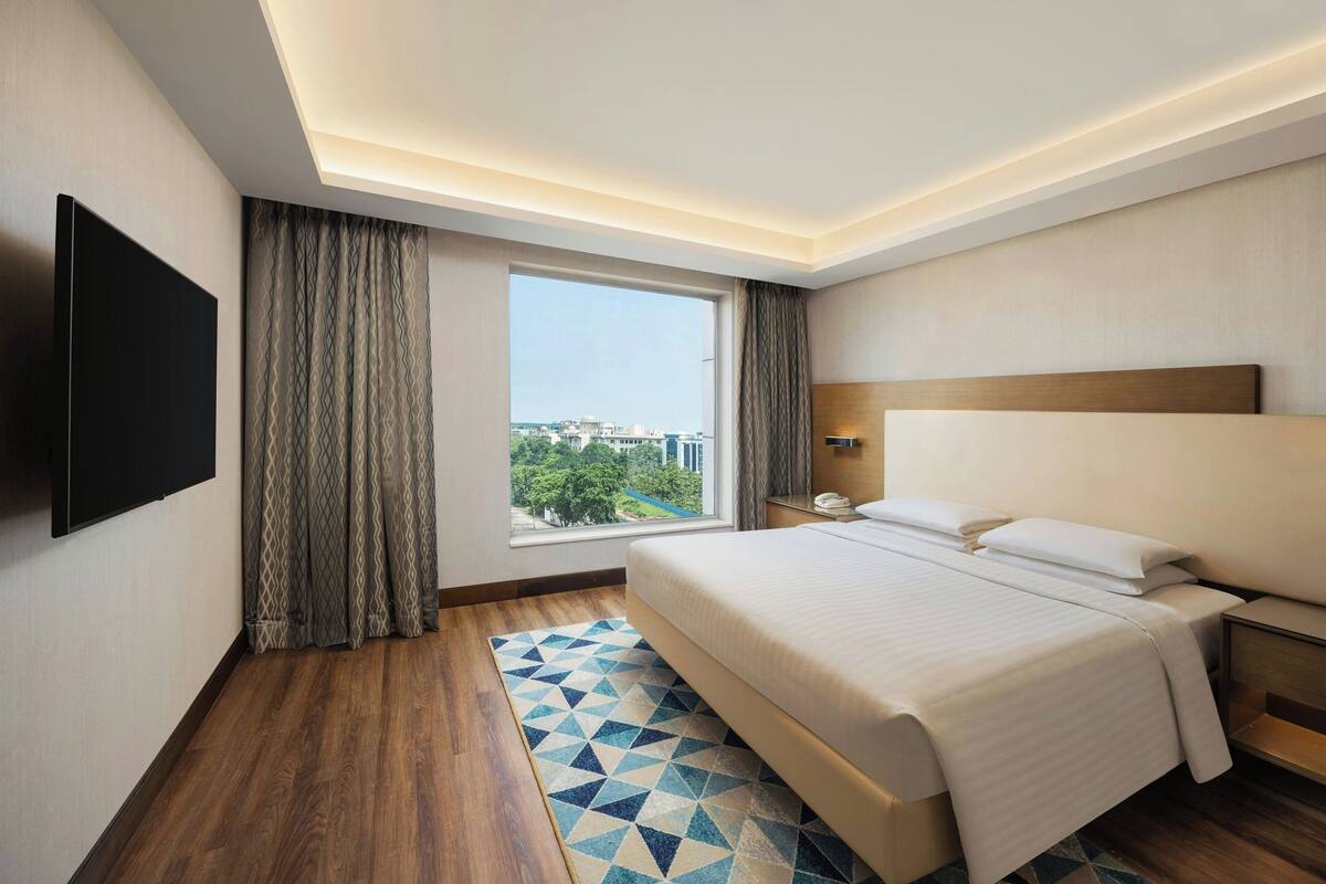 Habitación de hotel con una cama grande, un televisor de pantalla plana en la pared, una ventana con cortinas y vistas a la ciudad.