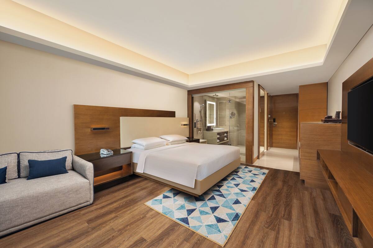 Habitación de hotel moderna con cama, sofá, televisión y baño visible a través de una puerta de cristal.