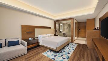 A modern hotel room with a bed, a sofa, a TV, and a bathroom visible through a glass door.