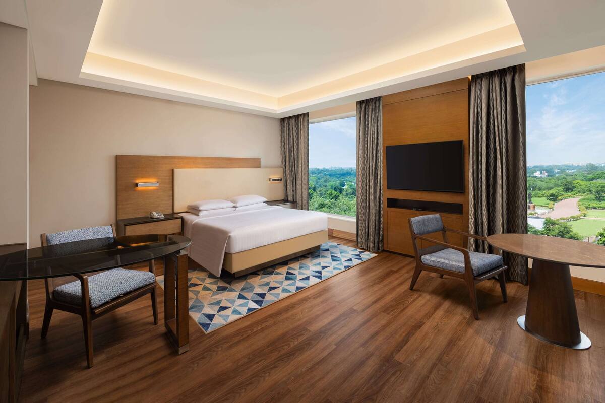Habitación de hotel con cama, escritorio, silla, televisor y un amplio ventanal con vistas.