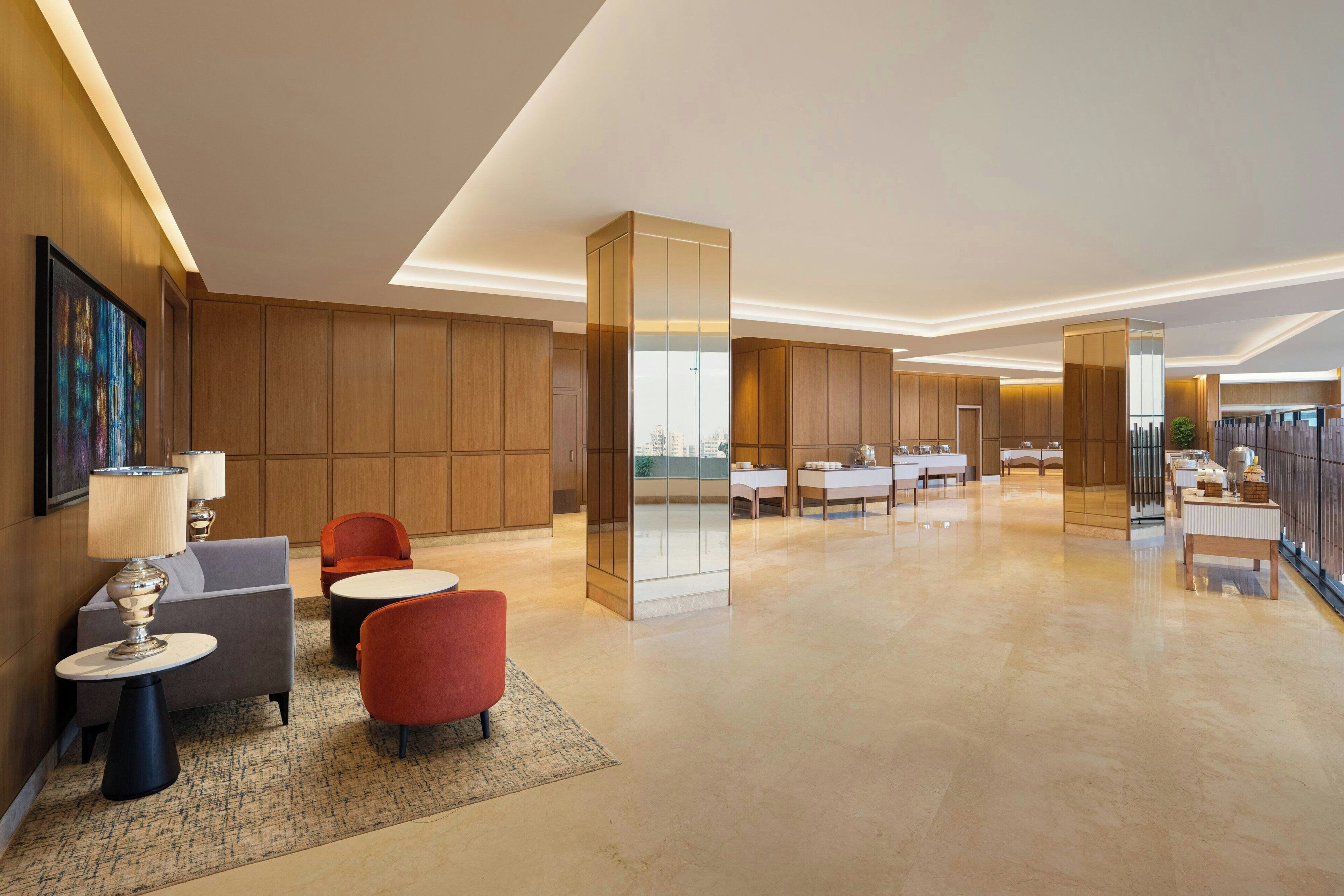 A modern hotel lobby with wooden paneling, a reception desk, seating area with a lamp, and a view of the city through large windows.
