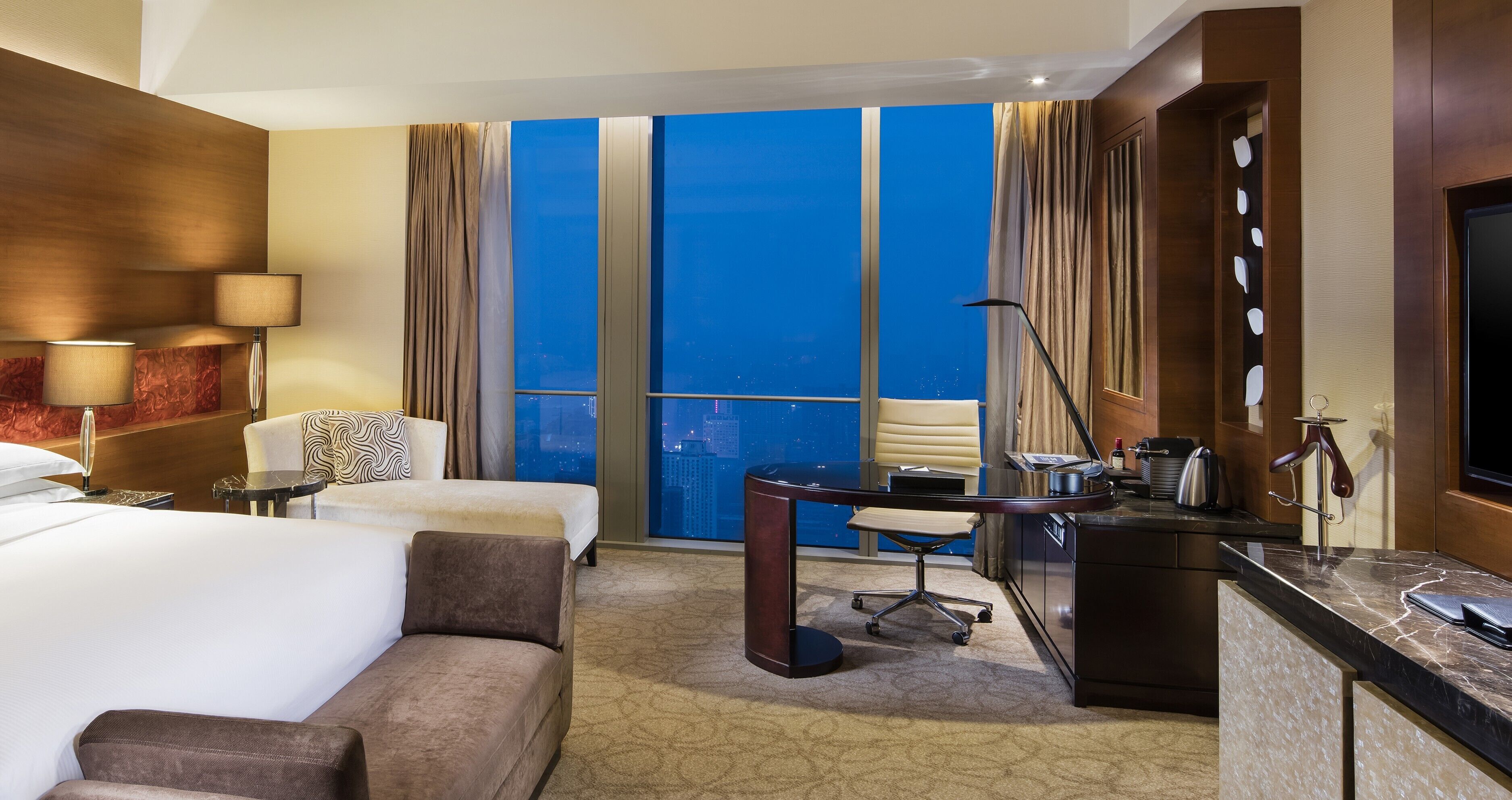 Une chambre d’hôtel équipée d’un lit, d’un bureau, d’une chaise, d’une télévision et d’une grande fenêtre donnant sur la ville.