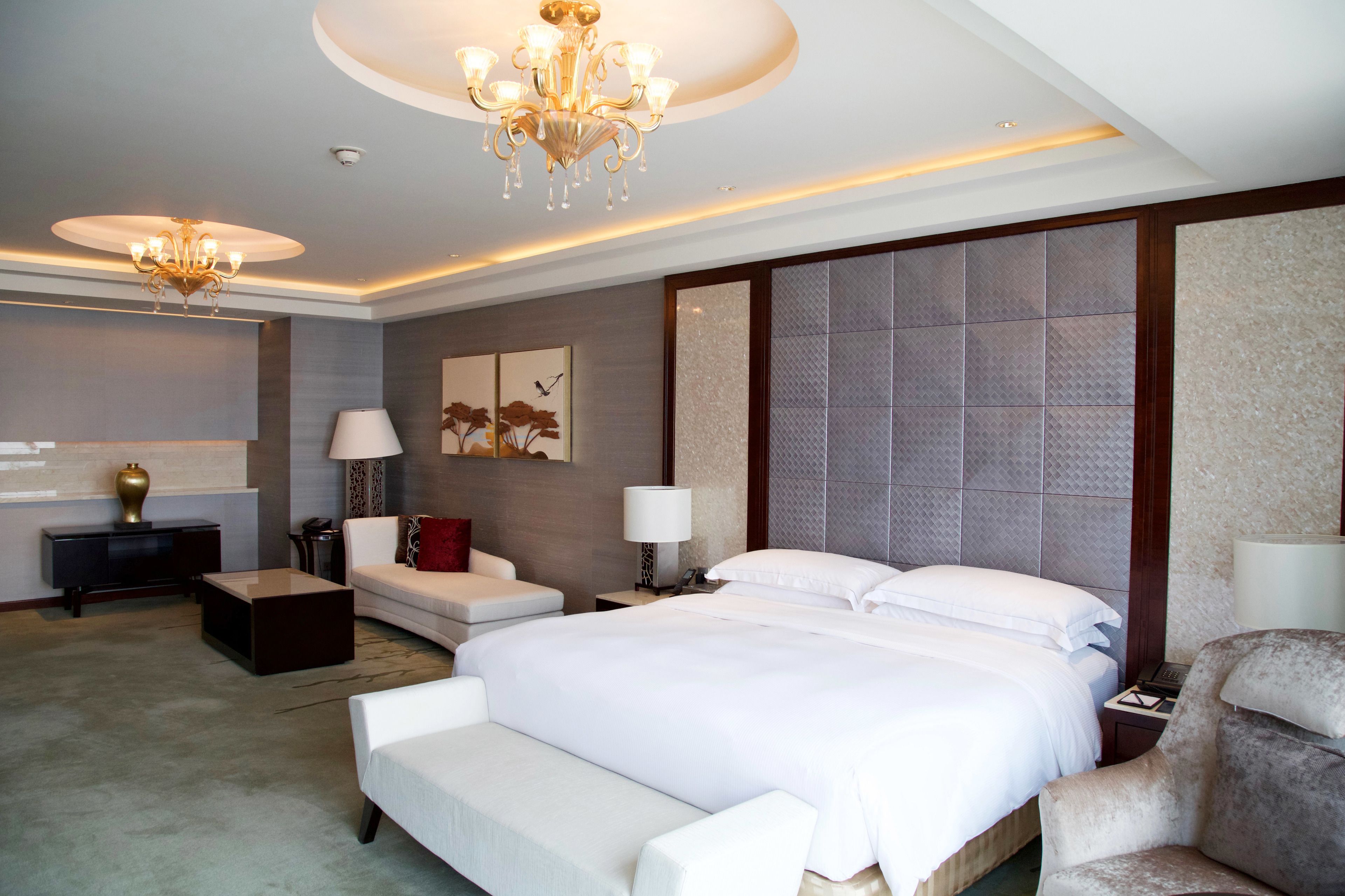 A hotel room with a large bed, a sitting area with a sofa and armchairs, a small table, and a decorative wall panel.