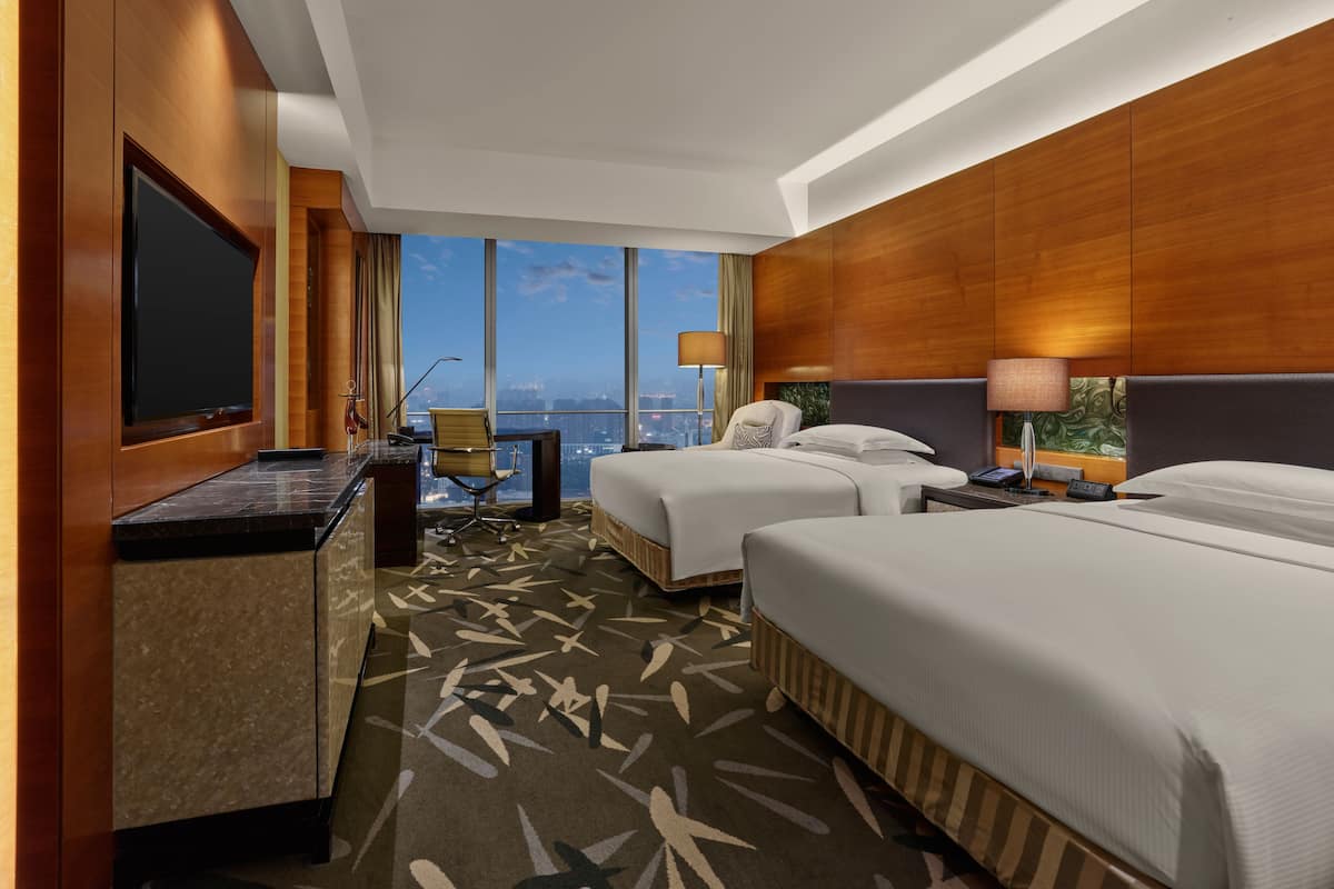 A hotel room with two beds, a desk with a flat-screen TV, a chair, a lamp, and a view of the cityscape.