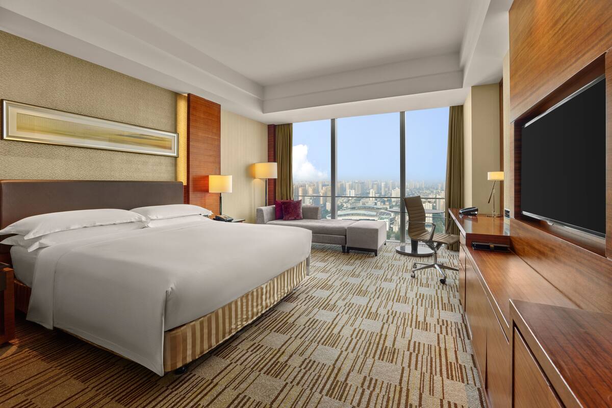 A hotel room with a large bed, a desk, a chair, a television, and a city view.
