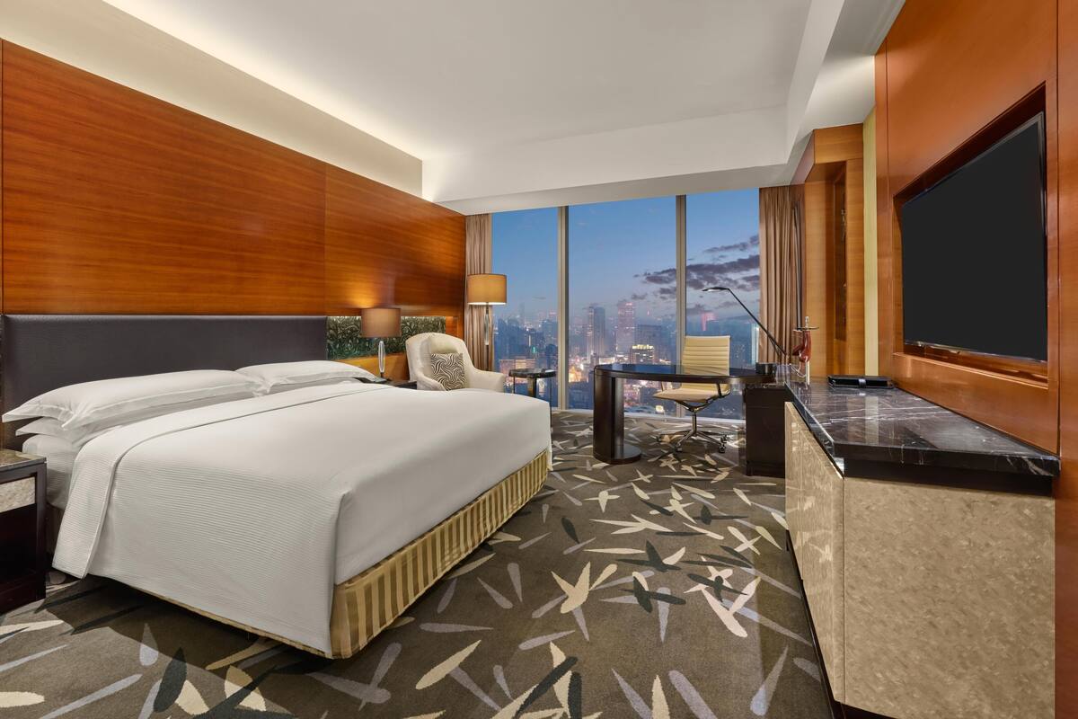 A hotel room with a bed, desk, chair, and a large window offering a city view.