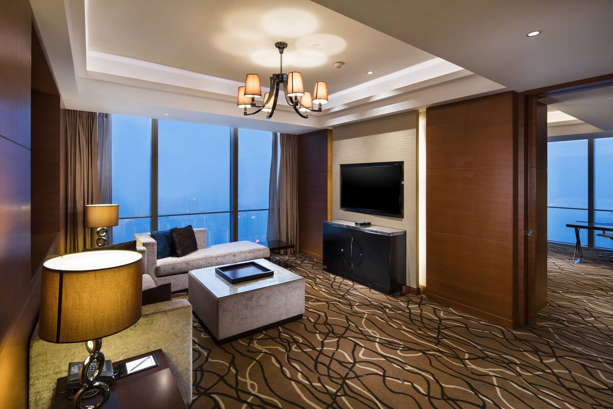 A hotel room with a large window, a sofa, a coffee table, a TV, and a chandelier.