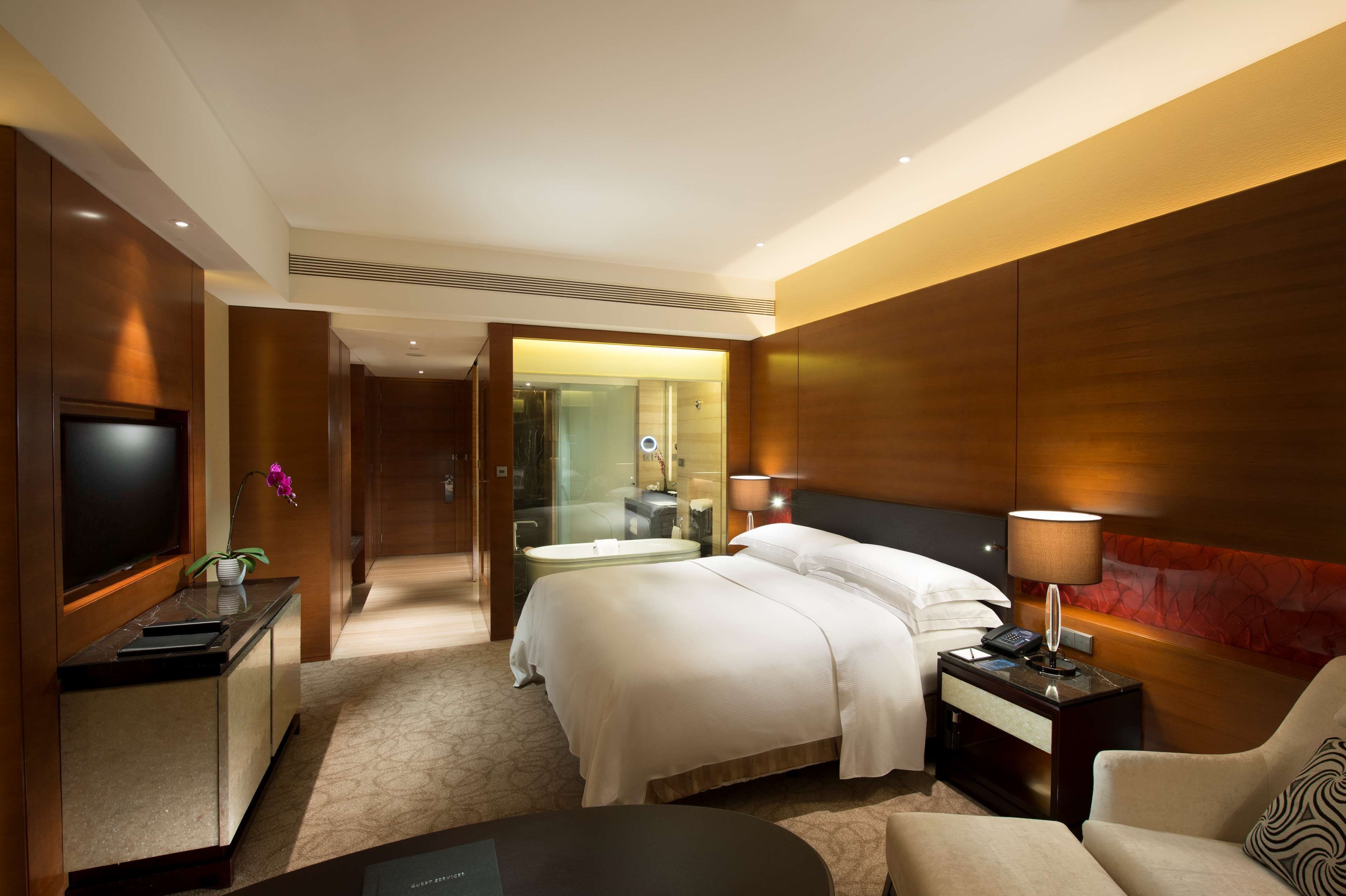 A hotel room with a bed, bedside table, lamp, TV, and a desk with a chair.