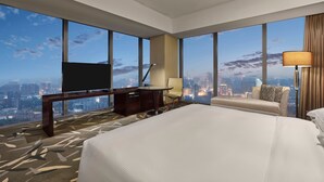 Habitación de hotel con una cama grande, un escritorio con televisor, una silla, una lámpara y vistas panorámicas de la ciudad a través de amplias ventanas.