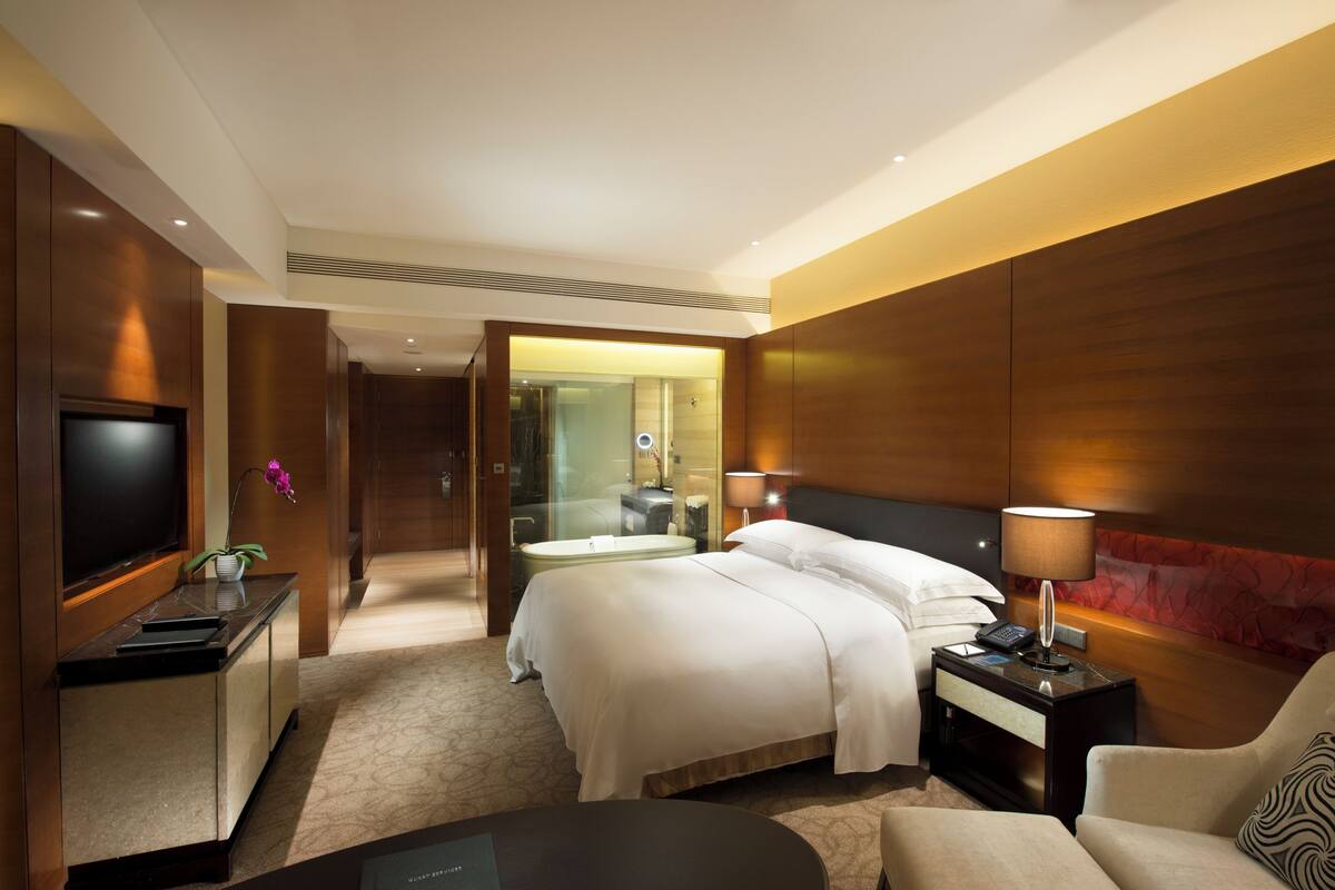 A hotel room with a bed, bedside table, lamp, TV, and a seating area.