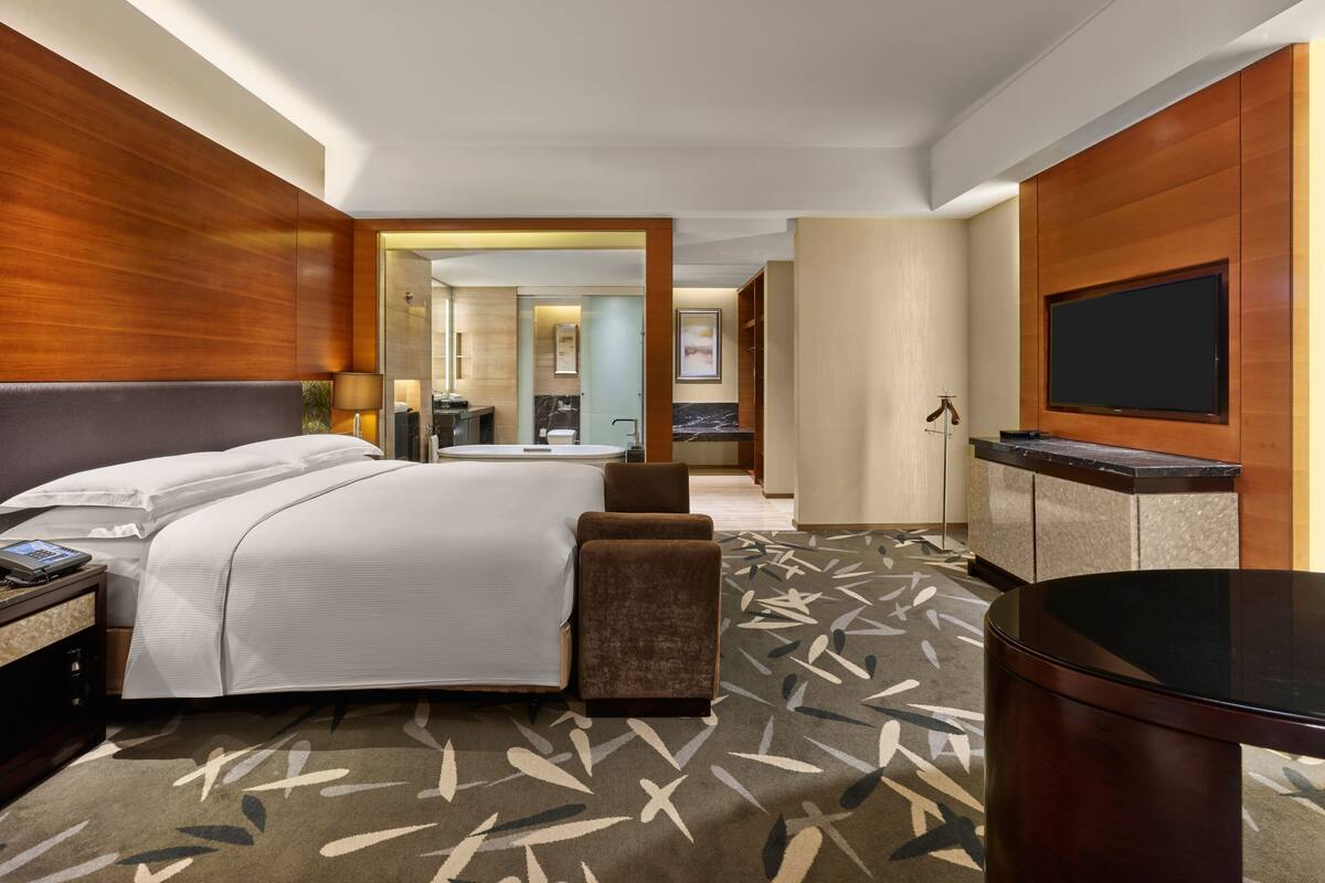 A hotel room with a large bed, a desk with a telephone, a flat-screen TV mounted on the wall, and a bathroom visible through an open door.