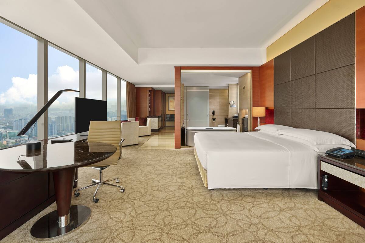 A hotel room with a large window, a desk, a bed, and a bathroom.