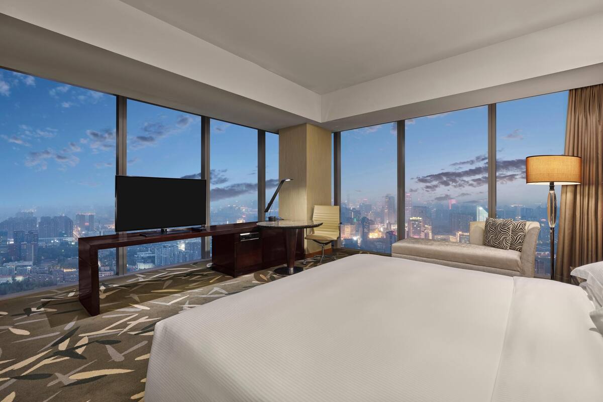 Habitación de hotel con una cama grande, un escritorio con televisor, una silla y vistas a la ciudad.