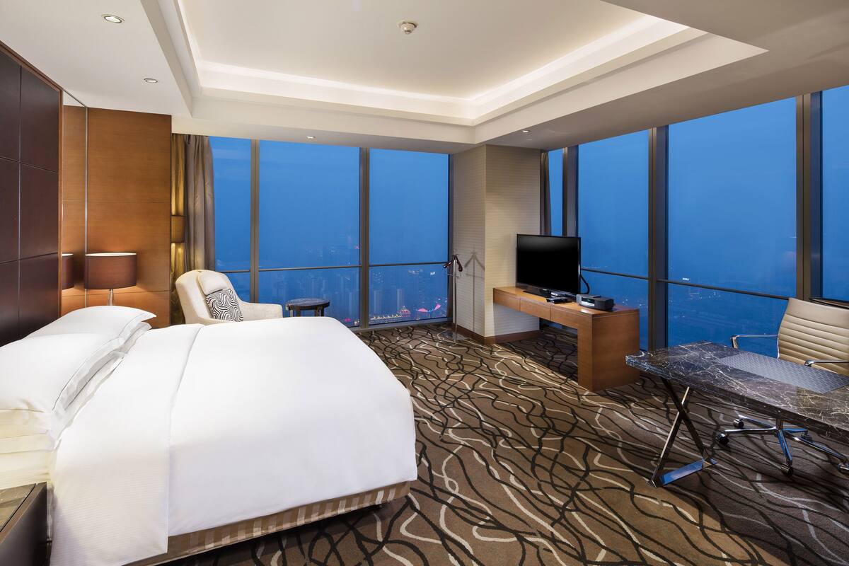 A hotel room with a large bed, a desk with a TV, a chair, and a view of the city through large windows.