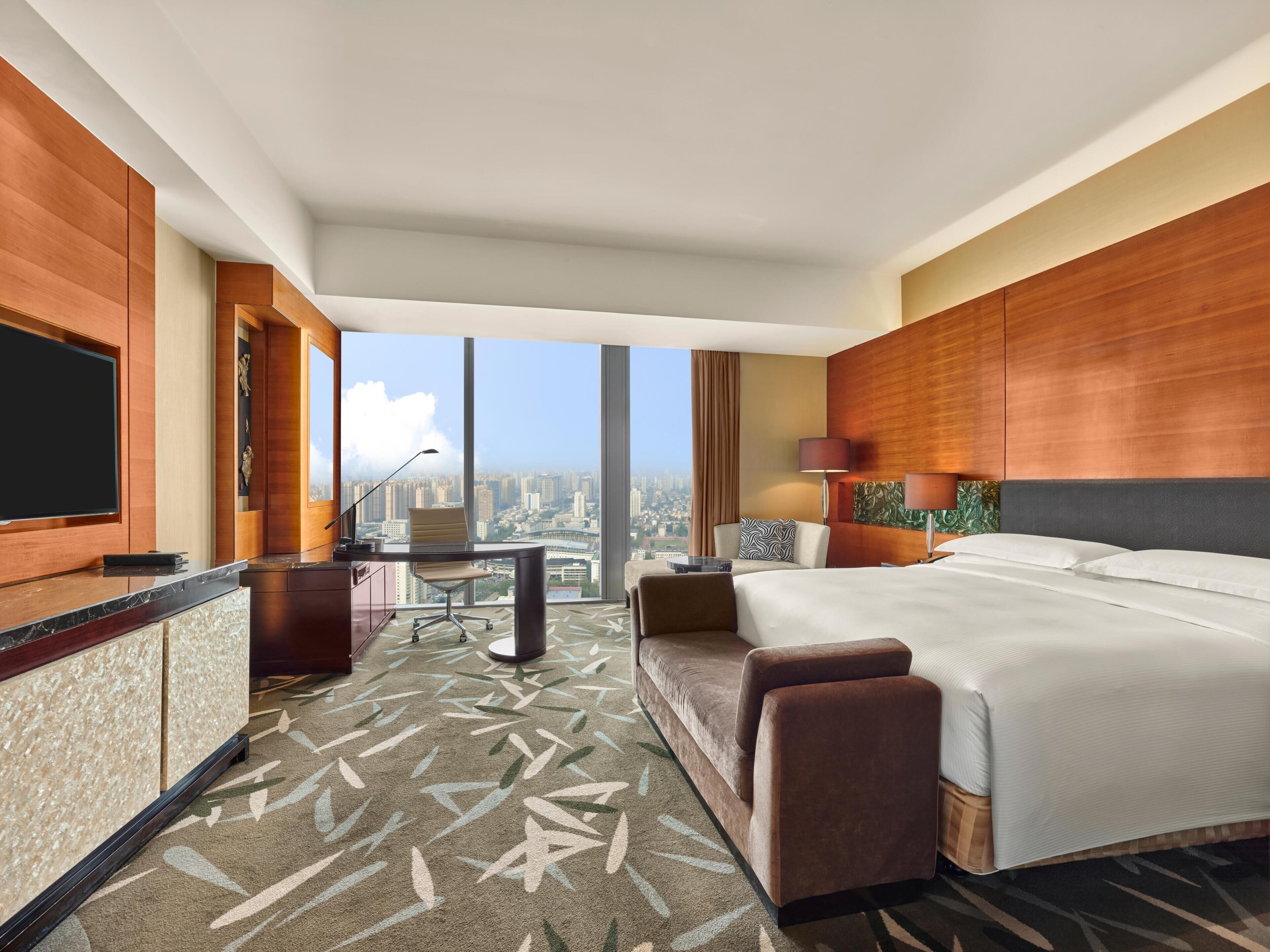 Ein Hotelzimmer mit einem großen Bett, einem Schreibtisch mit Stuhl, einem Fernseher und Blick auf die Stadt.