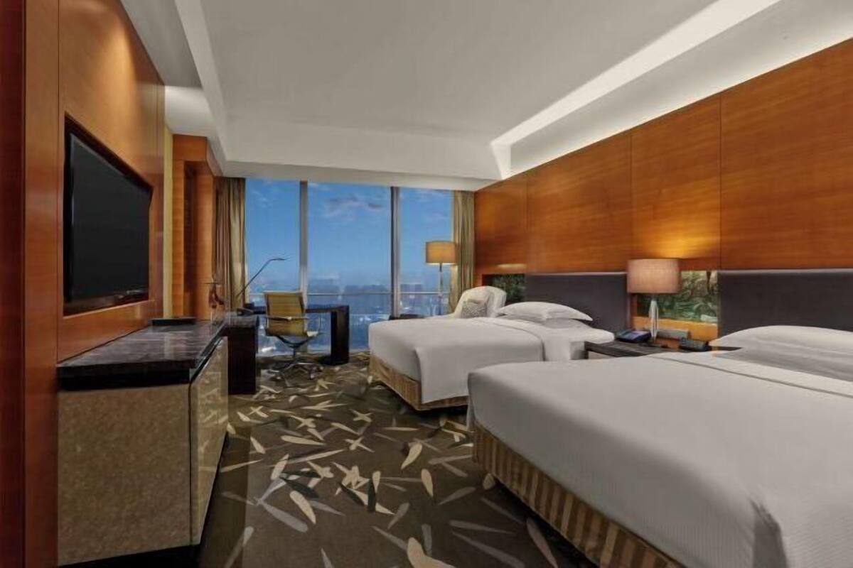 A hotel room with two beds, a TV, a desk, and a view of the city.