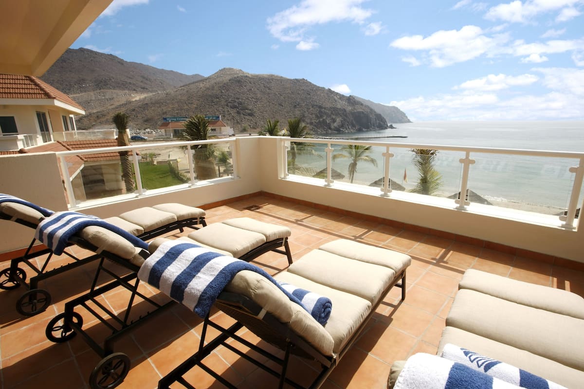 A balcony with sun loungers, a view of the sea, and a mountain in the background.