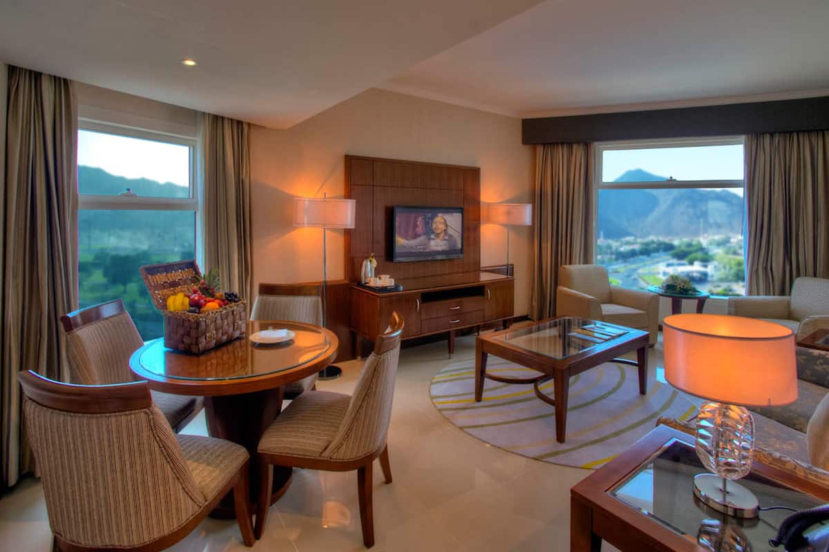 A hotel room with a dining area, a living room, a TV mounted on the wall, and large windows offering a mountain view.