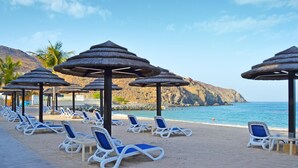 Une plage avec des transats et des parasols en chaume, un ciel dégagé et une côte rocheuse.