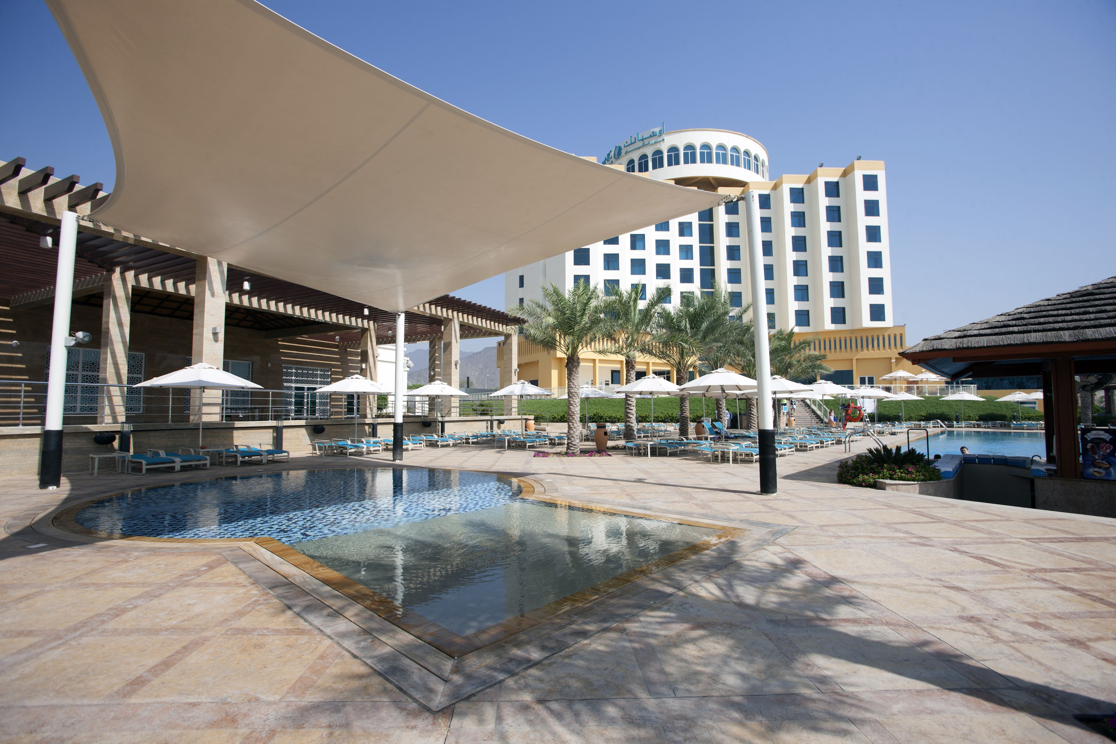 A hotel pool area with lounge chairs, umbrellas, and a large building in the background.