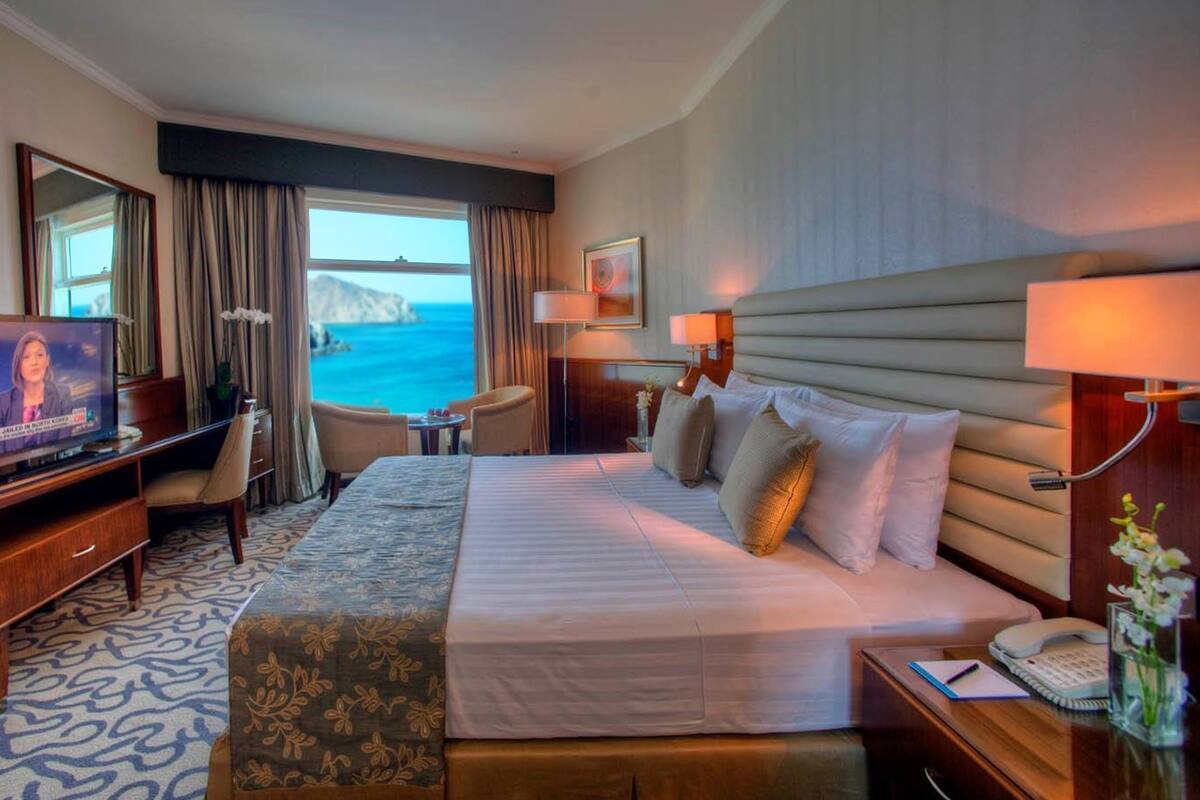 A hotel room with a bed, desk, TV, and a view of the sea.