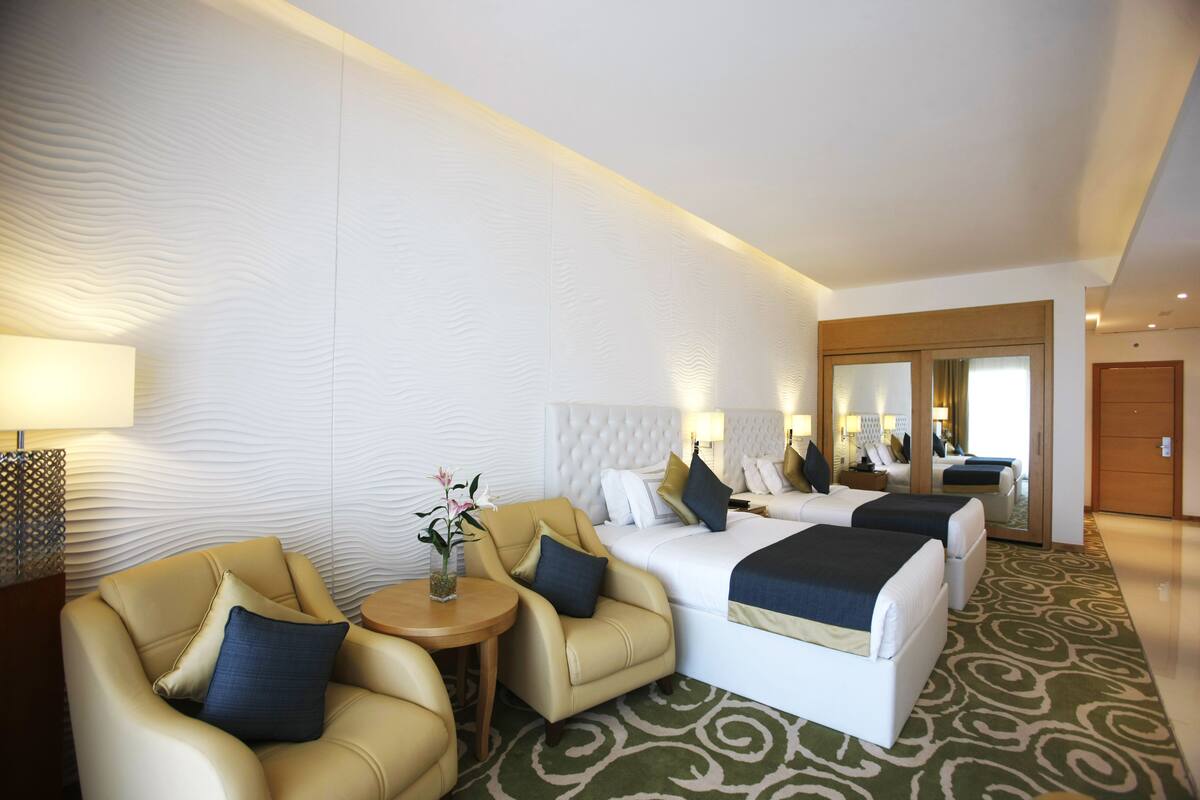 A hotel room with a large bed, two smaller beds, a sofa, a small table with a vase, and a mirror.