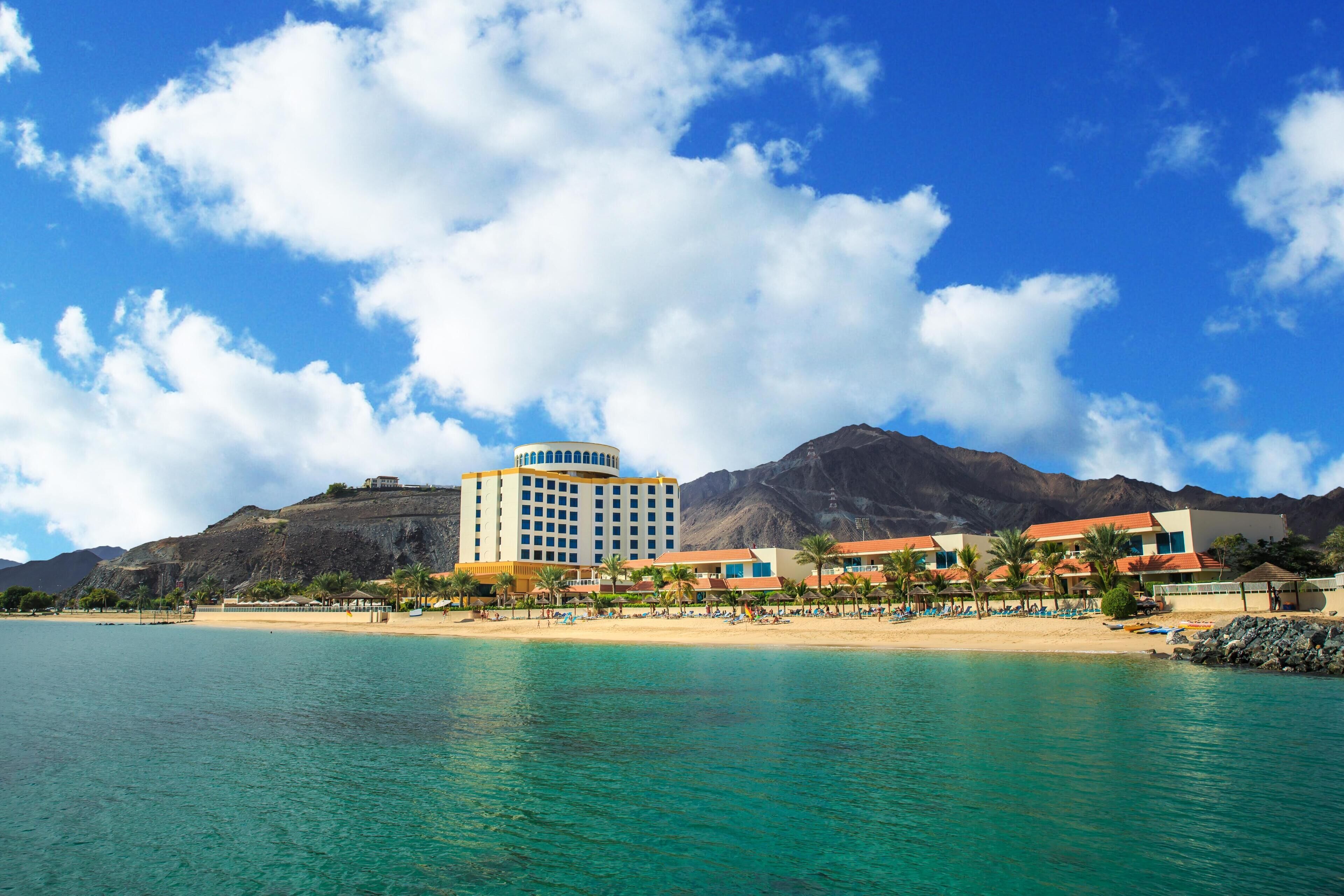 Un hotel sul lungomare con vista sulla spiaggia, un ampio edificio centrale e montagne sullo sfondo.