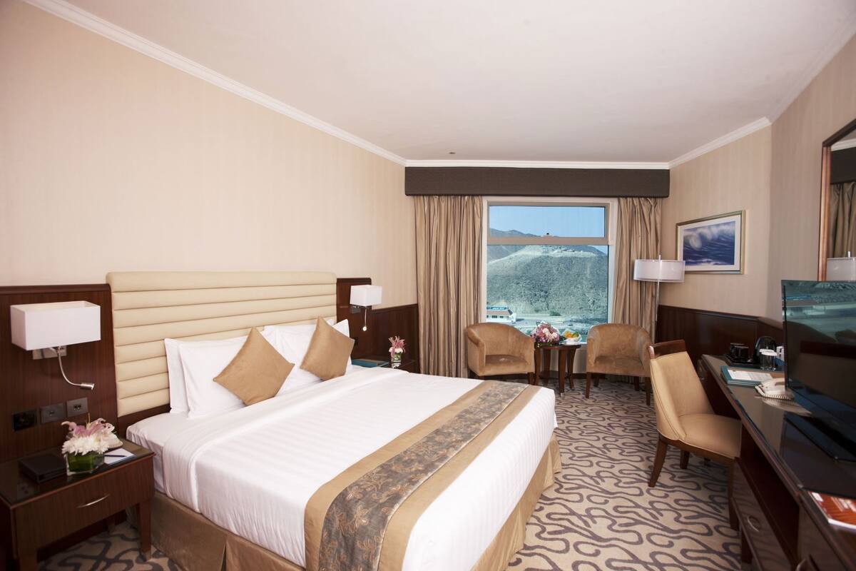 A hotel room with a large bed, two armchairs, a desk with a TV, and a window with a mountain view.