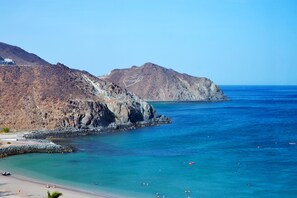 Paisaje costero con playa rocosa, mar azul transparente y montañas al fondo.