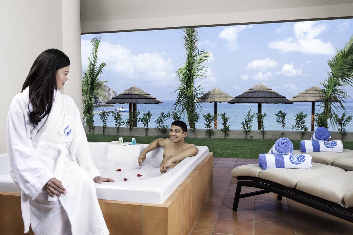 A room with a large bathtub, a sofa, and a view of the ocean with umbrellas.