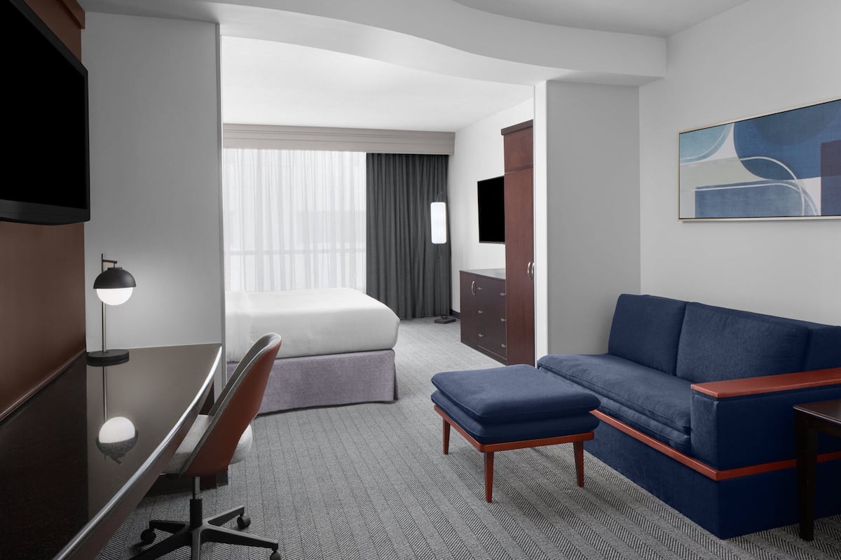 A hotel room with a bed, a desk, a chair, a sofa, a TV, and a painting on the wall.