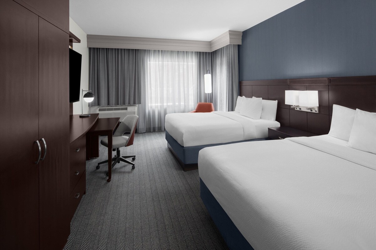 A hotel room with two beds, a desk with a chair, a TV, and a window with curtains.