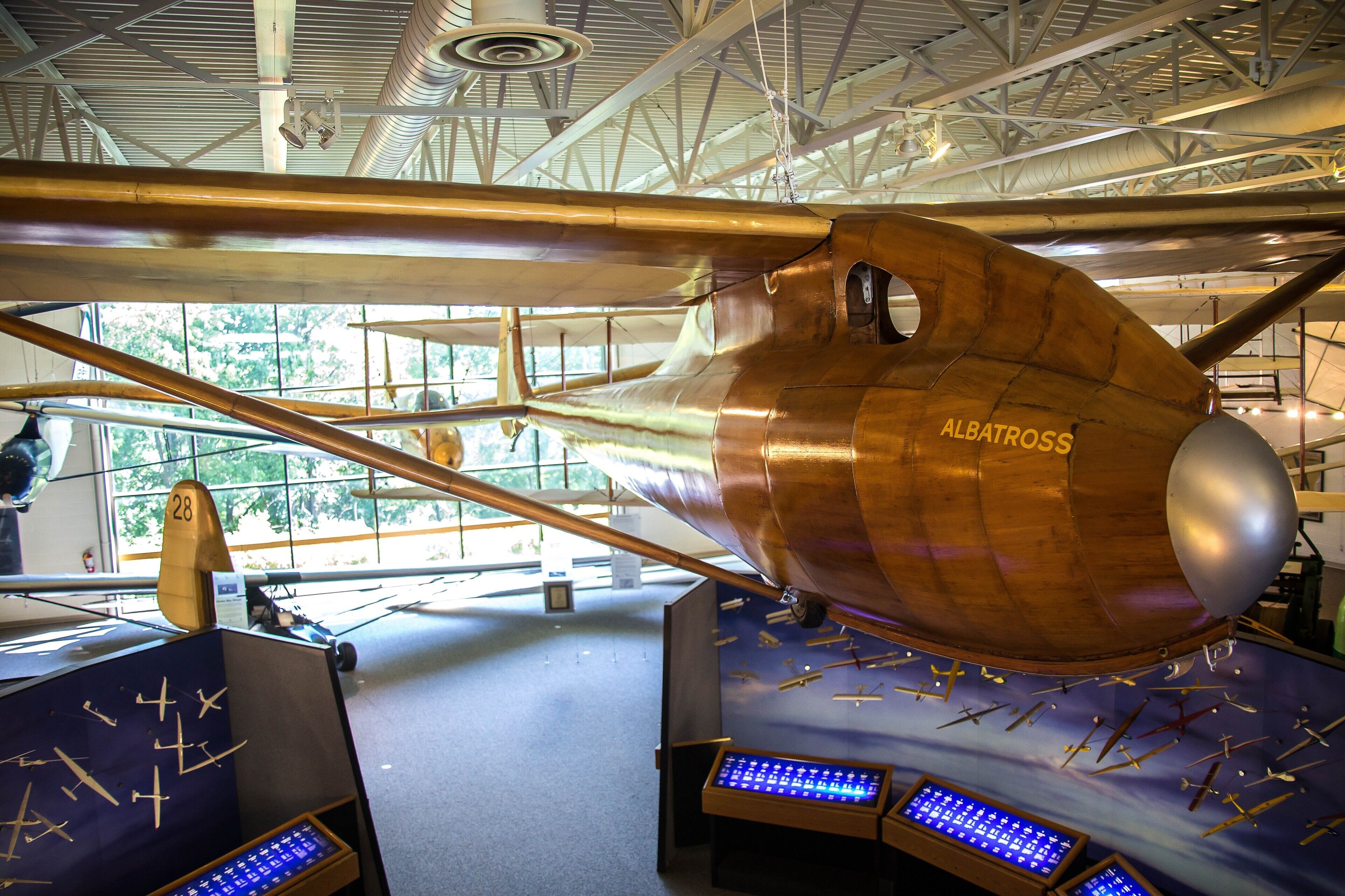 Una exposición de museo con una aeronave antigua que tiene el nombre "ALBATROSS" en el costado, rodeada de varios modelos de aviación y un panel de control.