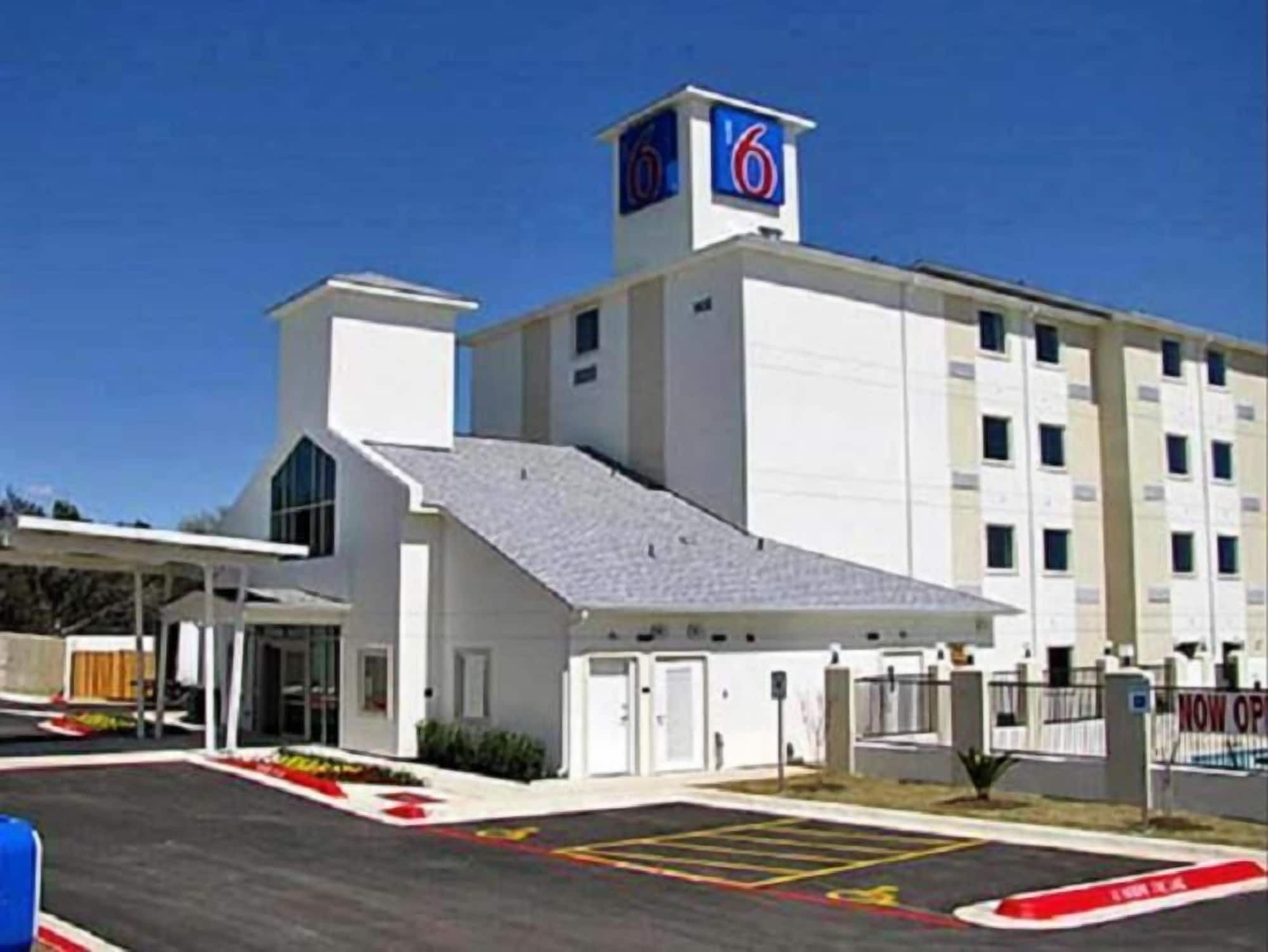 A modern hotel with a prominent '6' sign, a covered entrance, and a parking lot with marked spaces.