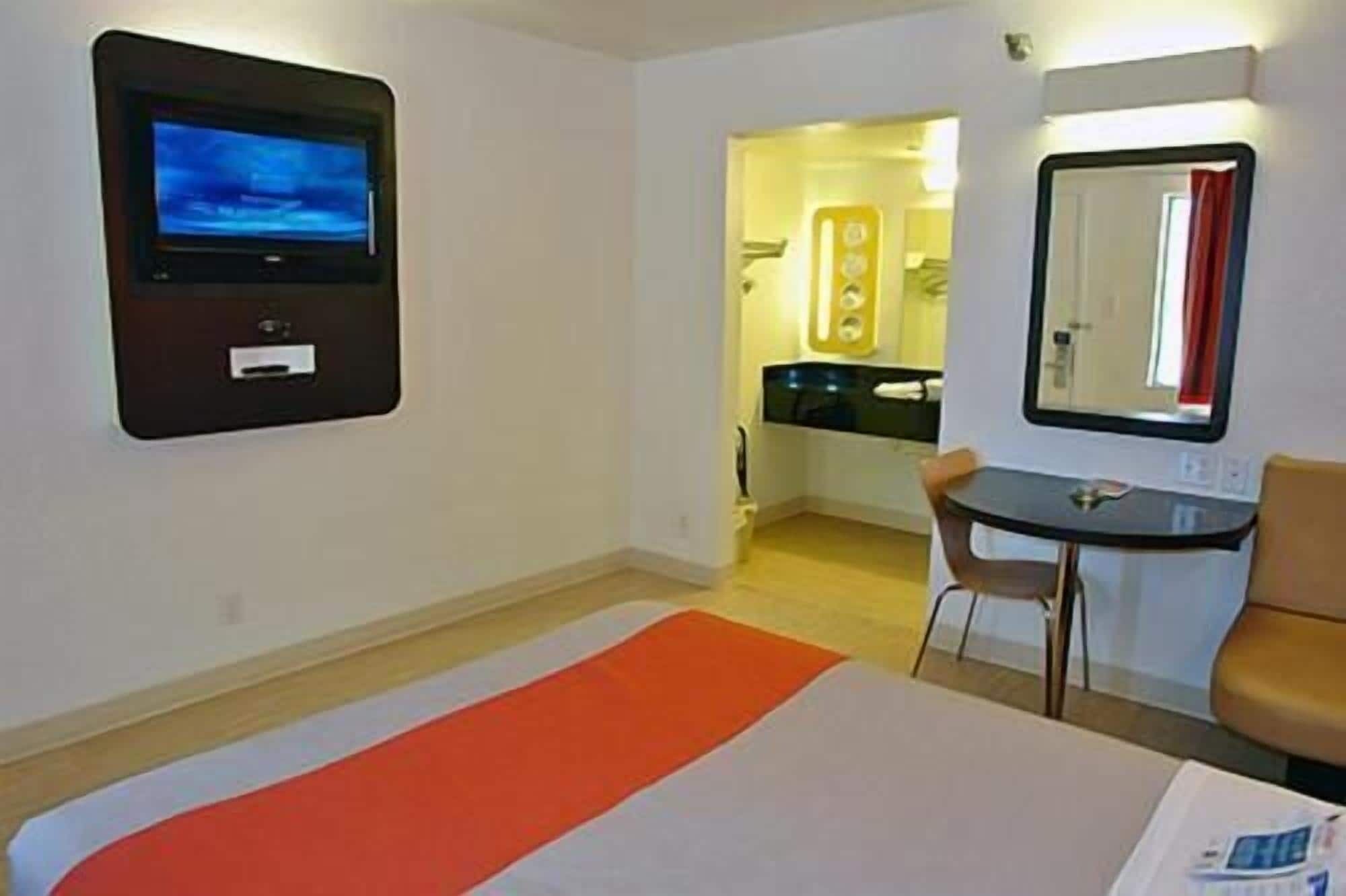 Habitación de hotel con una cama, televisión montada en la pared, una mesita con una silla y un espejo.