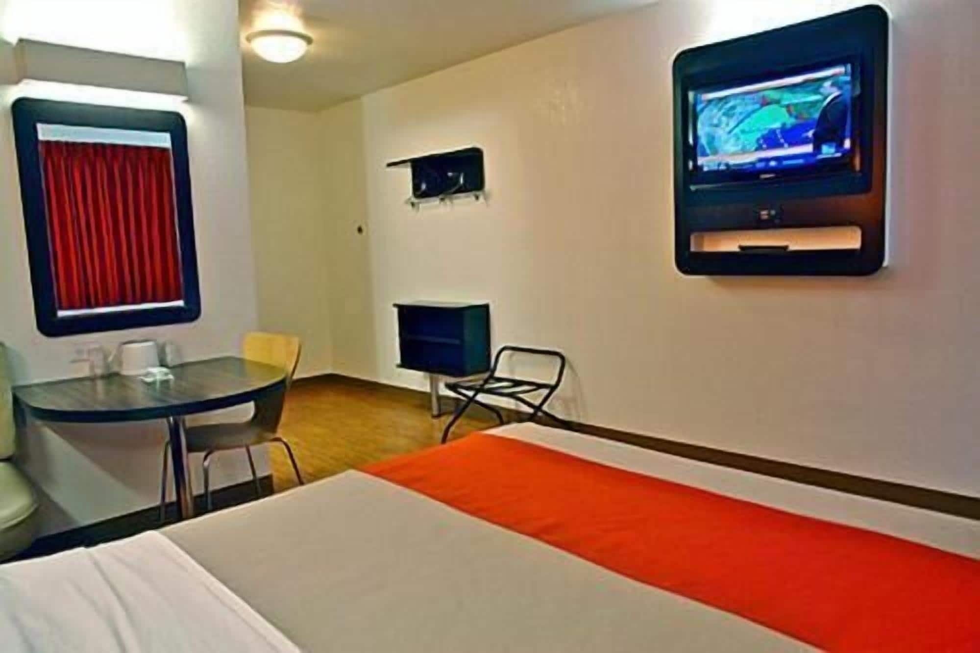 Habitación de hotel con cama, escritorio, silla, televisor montado en la pared y ventana con cortinas.