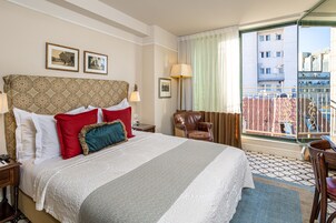 Habitación de hotel con una cama grande, una silla, un escritorio y un ventanal con vistas a edificios.