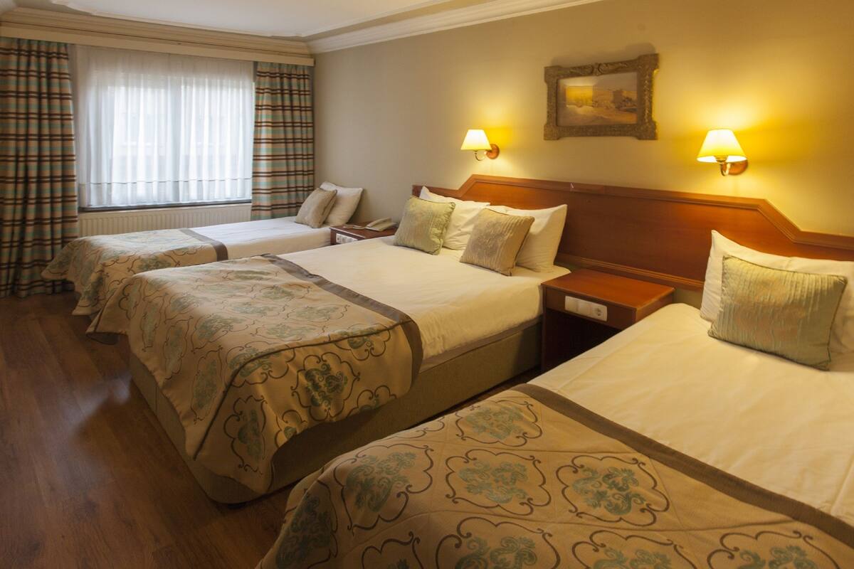 Een hotelkamer met drie bedden, elk met een houten hoofdbord, en een nachtkastje tussen de bedden. Aan weerszijden van de kamer hangen twee wandlampen.