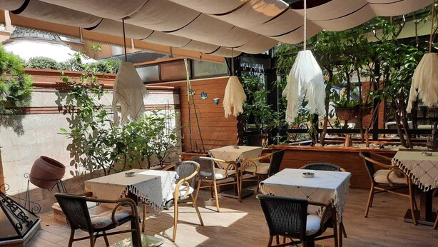 Un café al aire libre con mesas de madera y sillas de mimbre, rodeado de plantas en macetas y un muro de madera.